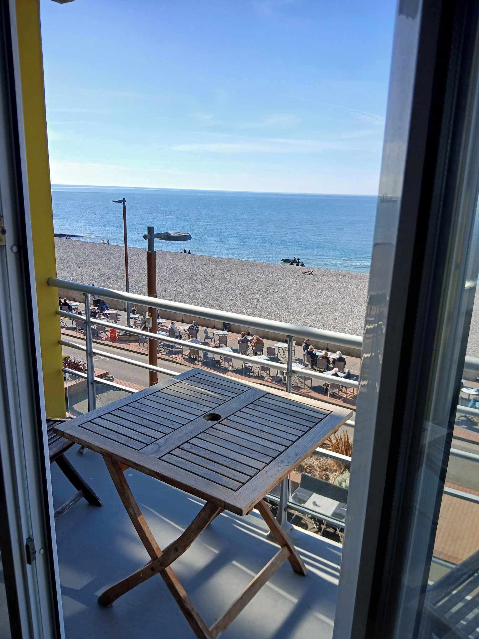 A balcony with a table and chairs, overlooking a beach with people and a clear sky.