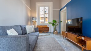 Een woonkamer met een grijze bank, een houten tv-meubel, een grote flatscreen-televisie, een kamerplant en een raam met uitzicht op gebouwen.