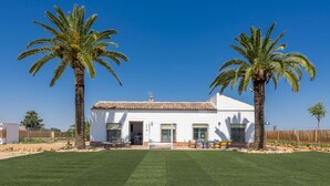 A white building with a tiled roof, two palm trees in front, and a well-maintained lawn. - Villa Colona, Los Algarbes J22g+94 (Los Algarbes- La Carlota)
