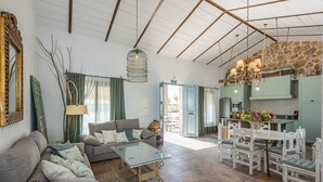 A spacious living area with a sofa, glass coffee table, and a dining table with chairs. A kitchen is visible in the background with a stone wall and a hanging light fixture. - Villa Colona, Los Algarbes J22g+94 (Los Algarbes- La Carlota)