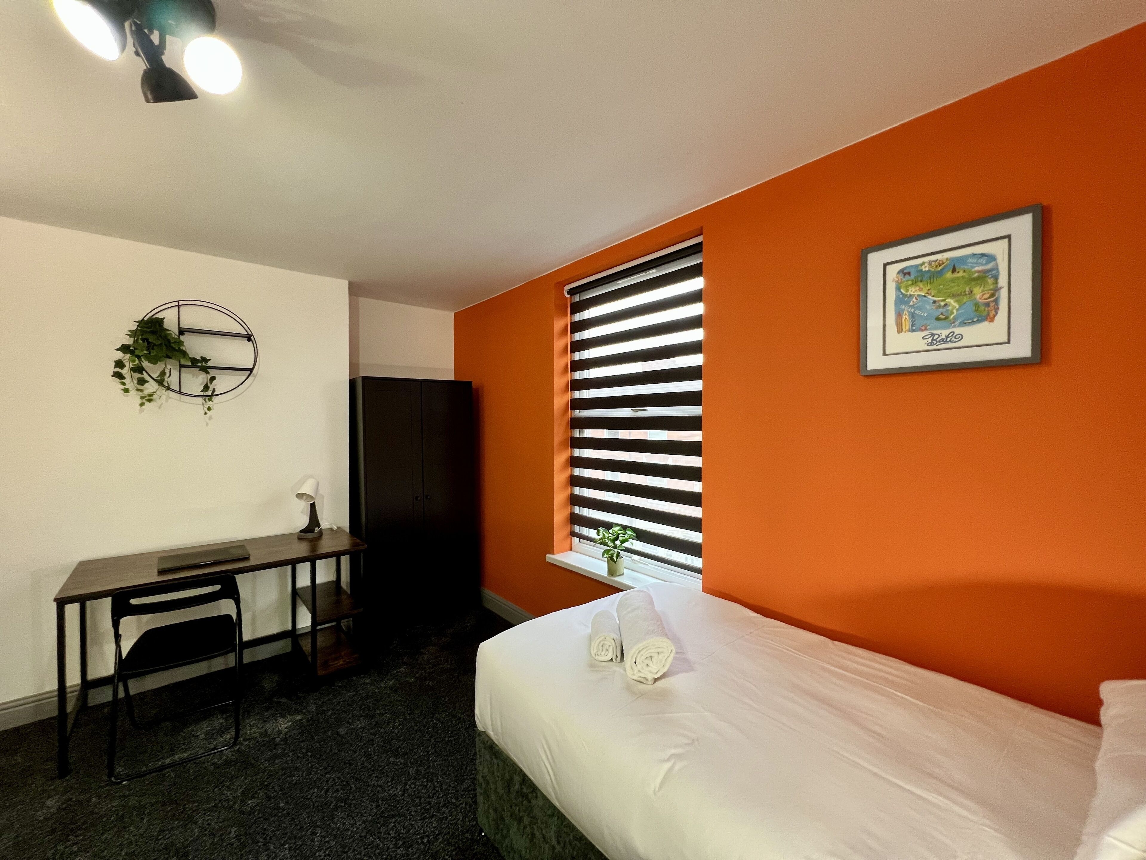 A room with a bed, a desk, a chair, a wardrobe, a window with blinds, a wall clock, and a framed map.
