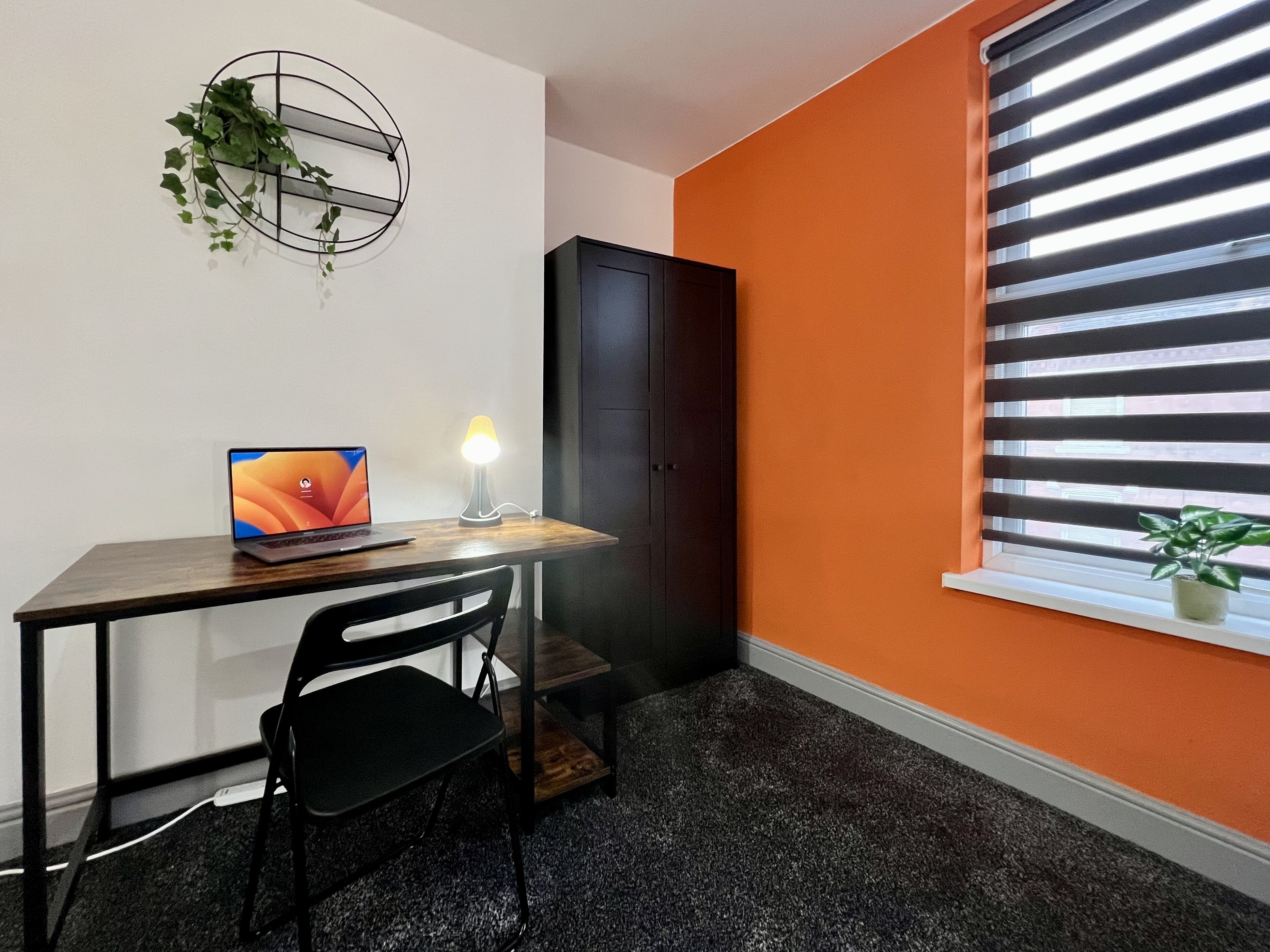Una habitación con escritorio, silla, ordenador portátil, lámpara y ventana con persianas.
