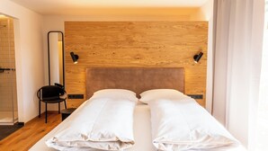 Une chambre d’hôtel avec une tête de lit en bois, deux oreillers blancs, une chaise noire et une cabine de douche en verre.