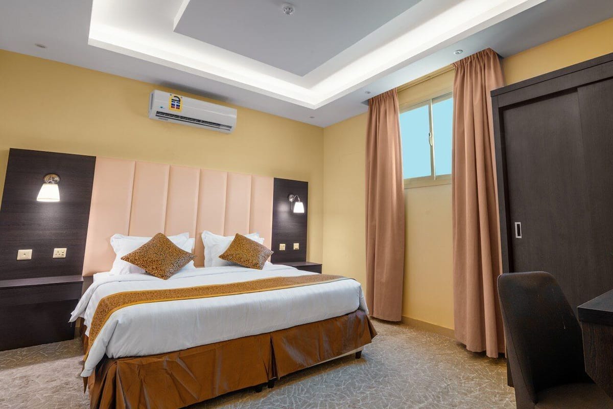 A hotel room with a bed, a desk, an air conditioning unit, and a window with curtains.