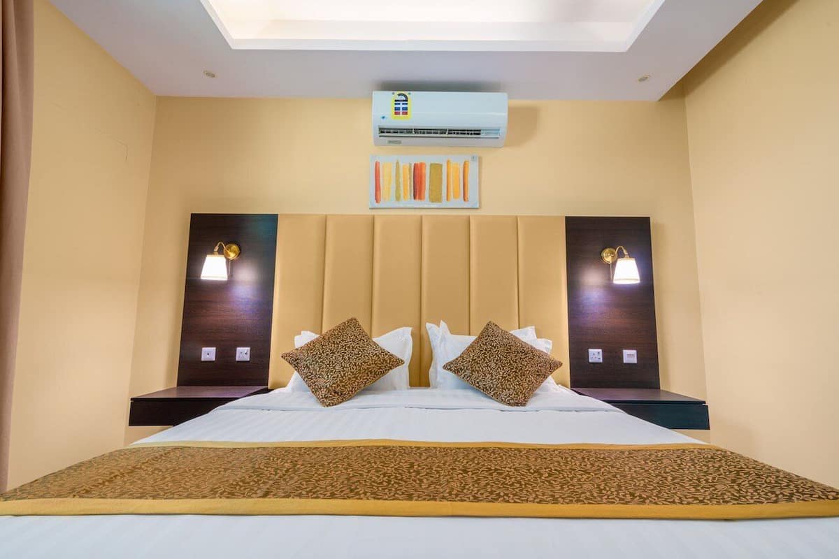 A hotel room with a bed, two wall-mounted lamps, an air conditioning unit, and a ceiling light.
