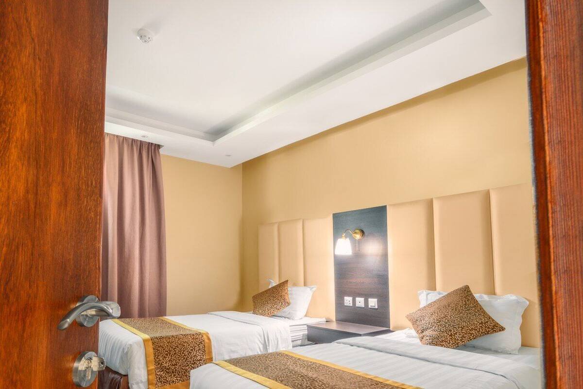 A hotel room with two beds, a wooden door, a beige wall, and a ceiling light.