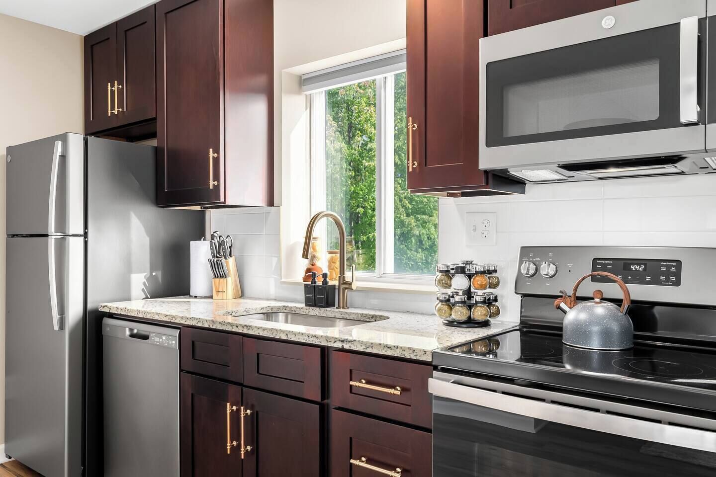 A modern kitchen with dark wood cabinets, stainless steel appliances, a granite countertop, and a window providing a view of greenery outside.