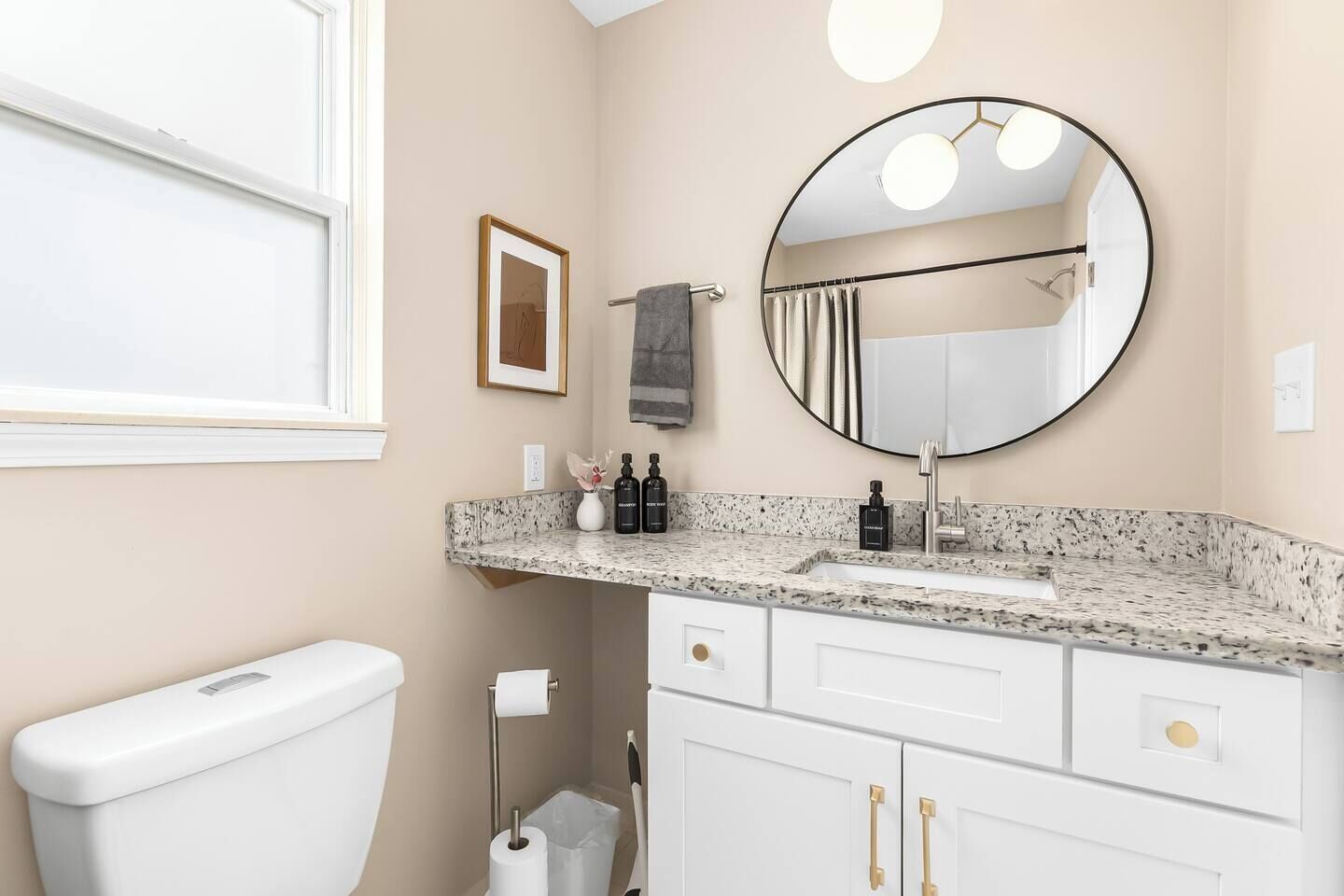 Ein Badezimmer mit Toilette, Waschbecken und Spiegel. An der Wand hängt ein gerahmtes Bild, eine Handtuchstange und ein Mülleimer sind vorhanden.