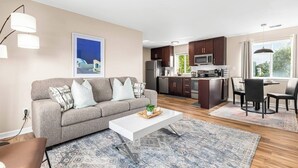 Ein modernes Wohnzimmer mit grauem Sofa, weißem Couchtisch und einem gemusterten Teppich. Eine Küche mit dunklen Schränken sowie ein Essbereich mit Tisch und Stühlen sind zu sehen.