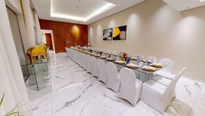 A long dining table with white chairs, set for a formal event, in a well-lit room with marble flooring and a wooden accent wall. - Suknai Resort (Riyadh)
