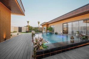 A modern outdoor pool area with a wooden deck, surrounded by a low wall with a climbing plant. - Suknai Resort (Riyadh)