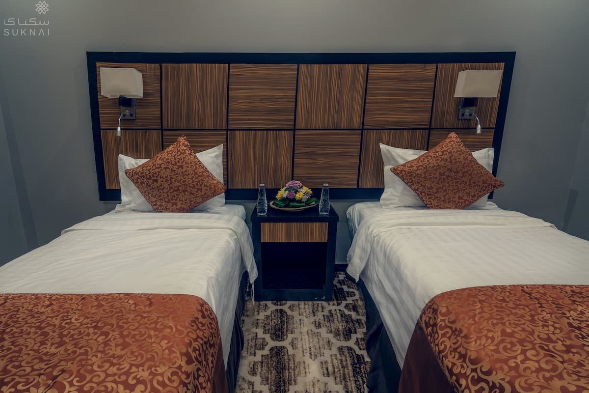 Habitación de hotel con dos camas, cabecero de madera, ropa de cama blanca, almohadas estampadas, una mesita con un florero y una alfombra decorativa.