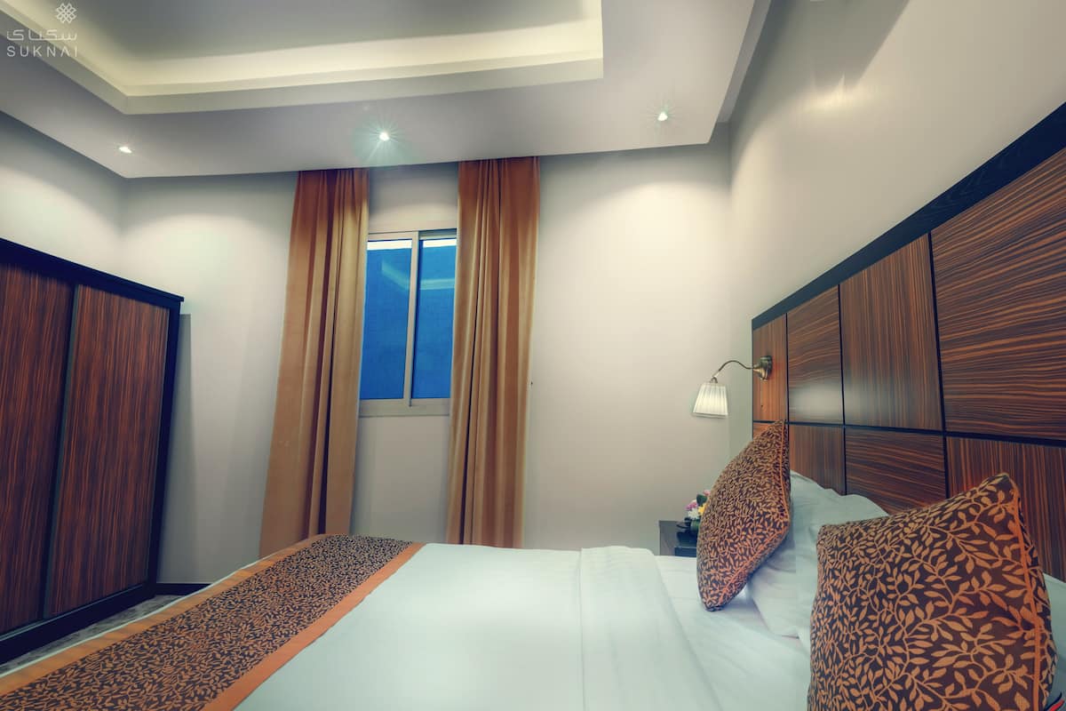 Habitación de hotel con una cama grande, cabecera de madera, una mesita de noche con lámpara y una ventana con cortinas.