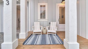 A hallway with white painted columns, a blue striped rug, two white Adirondack chairs, and a small decorative vase.