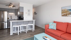 A modern kitchen with white cabinets, a stainless steel refrigerator, and a dining area with white stools. - Oceanfront: Beach, Sand and Views at Coral Reef! (North Topsail Beach)