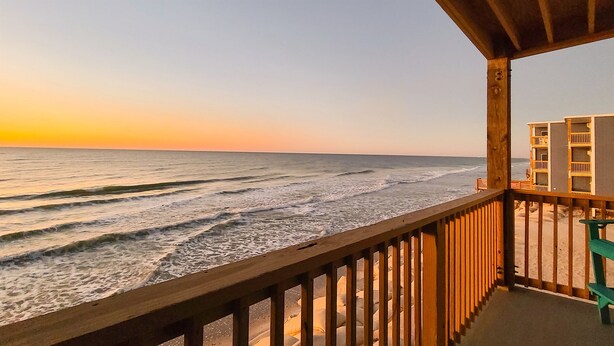 A wooden balcony overlooking the ocean with waves, a sandy beach, and a building with multiple balconies in the background. - Oceanfront: Beach, Sand and Views at Coral Reef! (North Topsail Beach)