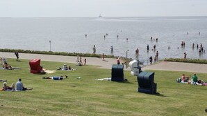 Ett grönområde där flera personer sitter och kopplar av och några promenerar längs vattnet.