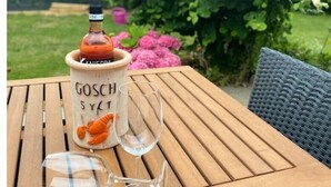 En flaska Gosch Syt-vin, två vinglas och en plastpåse på ett träbord utomhus.