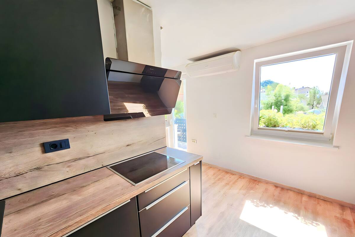 A modern kitchen with a built-in stove, a window with a view of greenery, and wooden flooring.