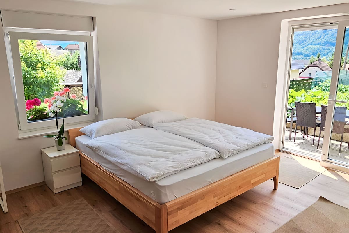 A bedroom with a wooden bed, a nightstand, a window with flowers, and a view of a garden and buildings.
