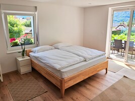 A bedroom with a wooden bed, a nightstand, a window with flowers, and a view of a garden and buildings.