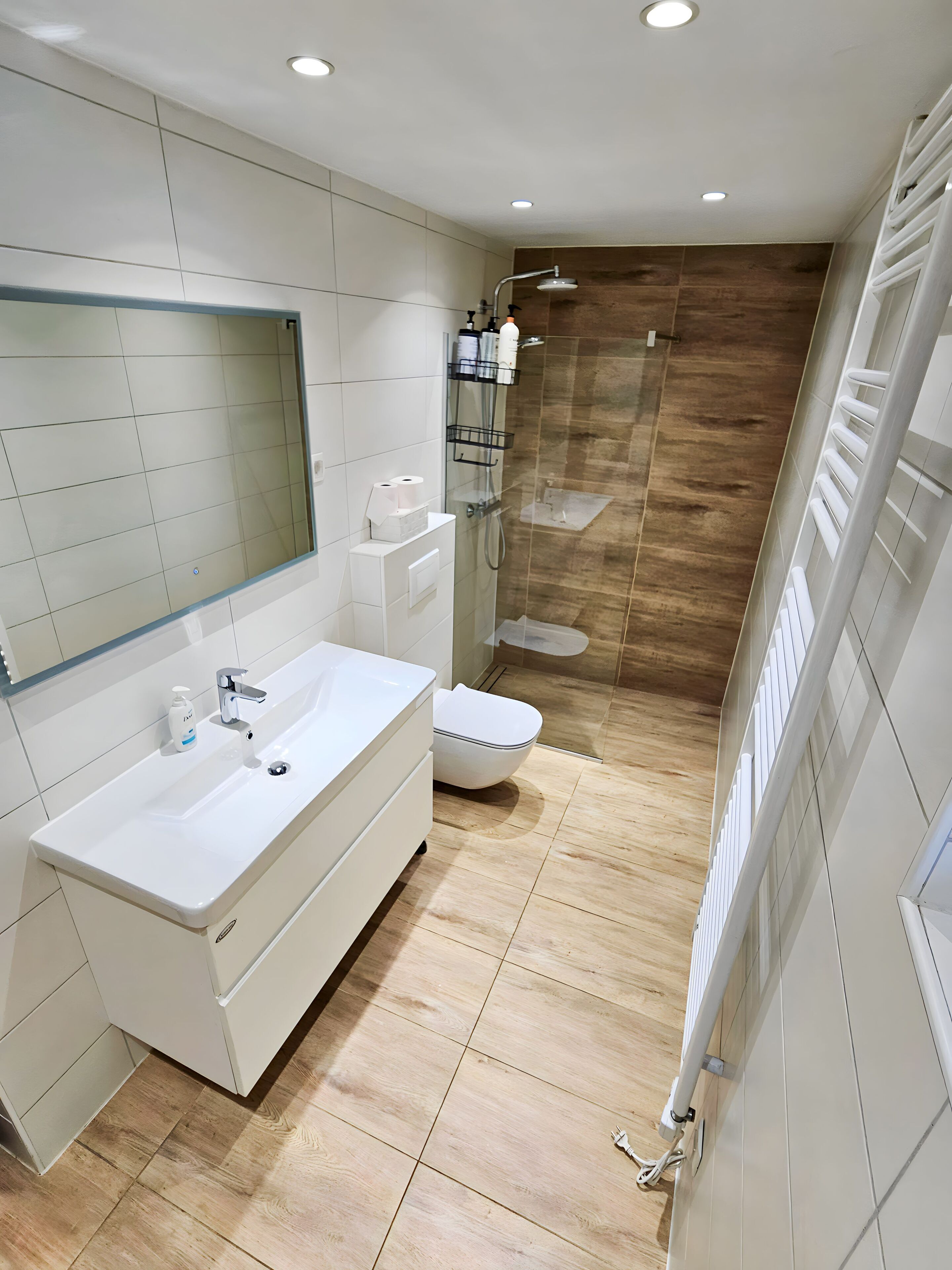 Un baño moderno con tocador blanco, un espejo grande, ducha con puerta de vidrio y suelo de madera.