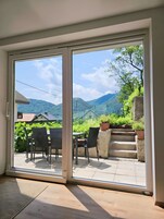 A room with a sliding glass door leading to an outdoor patio with a table and chairs, surrounded by potted plants and a view of a mountain landscape.