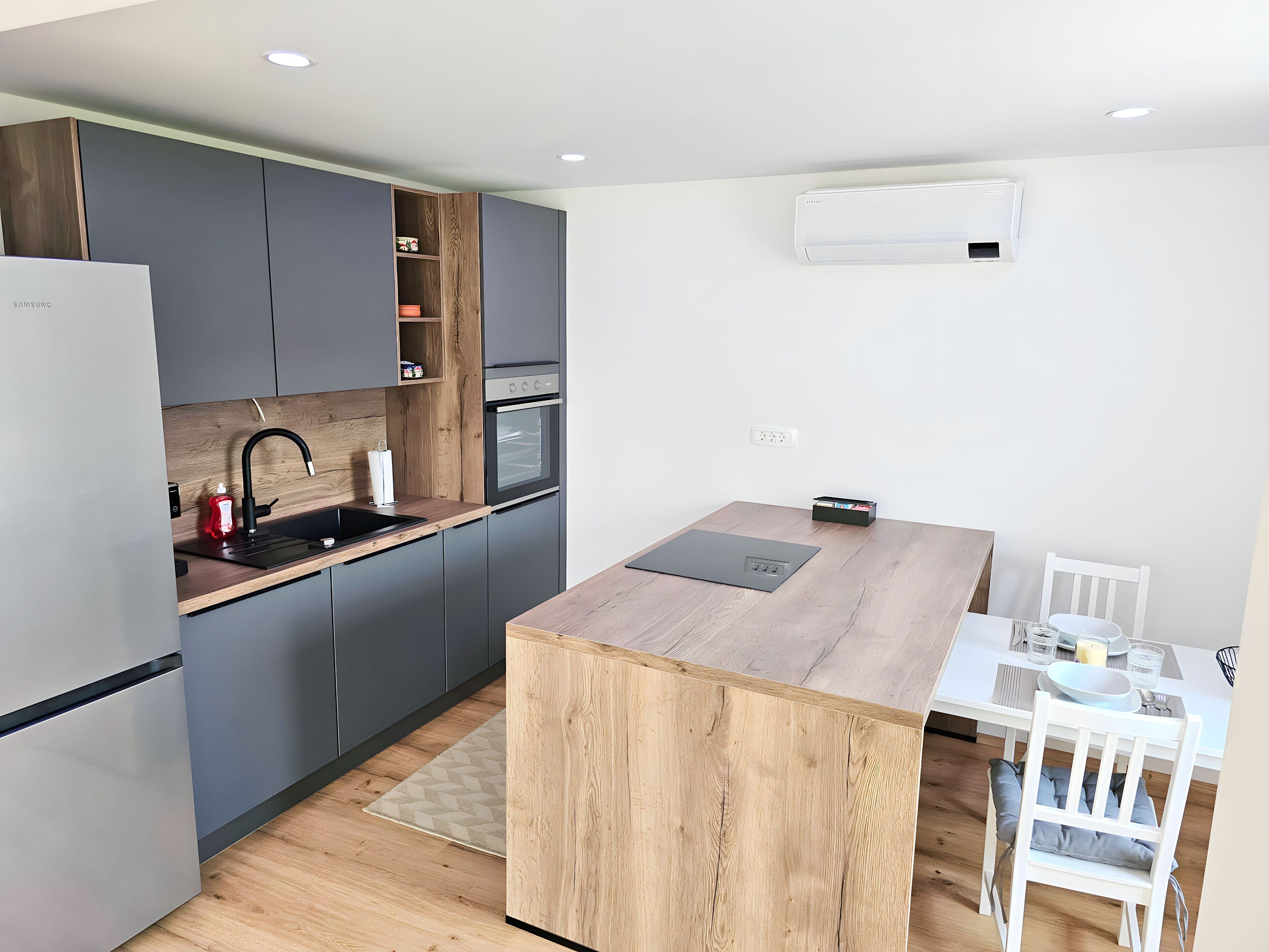 A modern kitchen with grey cabinets, a wooden countertop, a built-in oven, and a white dining table with chairs.