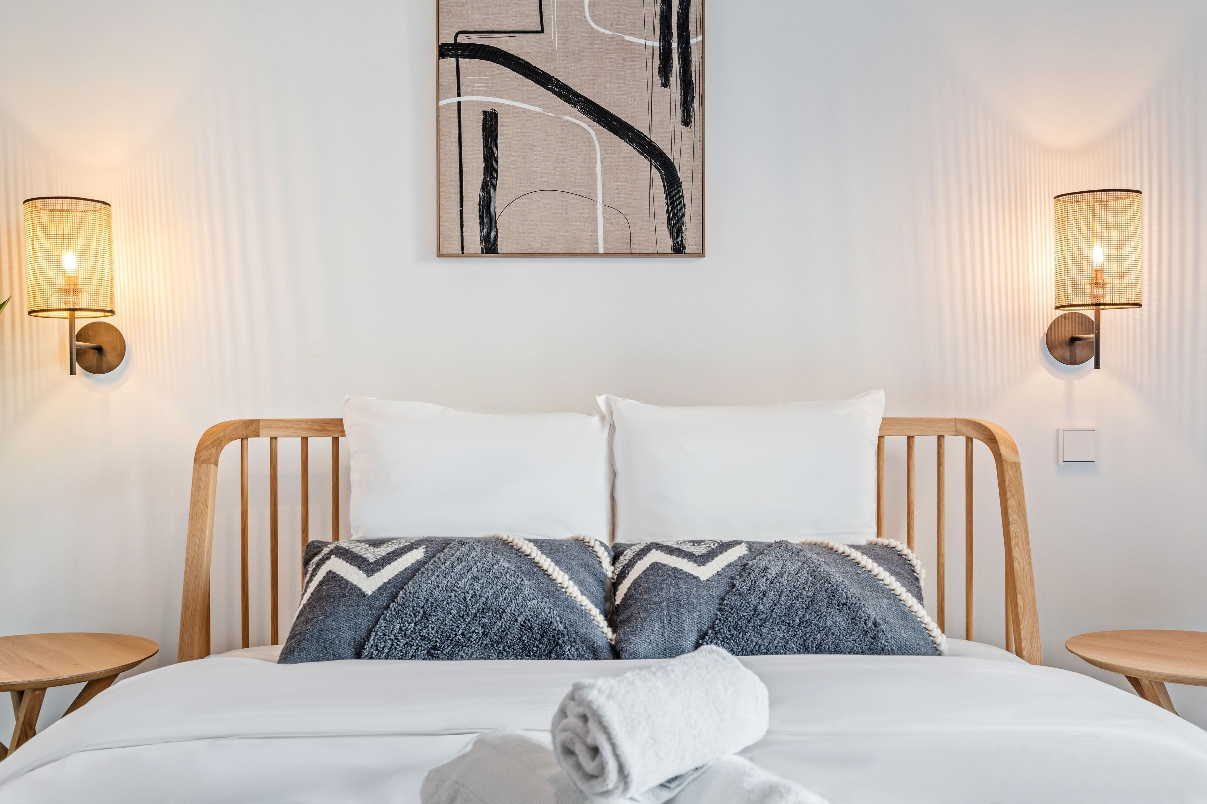 A neatly made bed with a white duvet and two pillows, a rolled towel on the bed, and two wall-mounted lamps on either side.