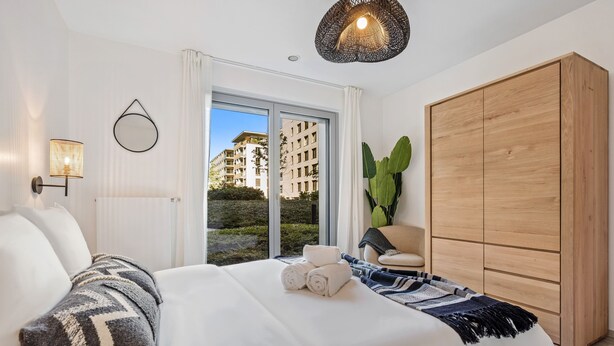 Un dormitorio moderno con una cama grande, un armario de madera, una planta en maceta y vistas a edificios a través de un ventanal.