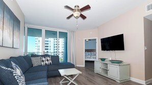 A modern living room with a sectional sofa, a small white table, a ceiling fan, a TV mounted on the wall, and a view of the city through large windows.