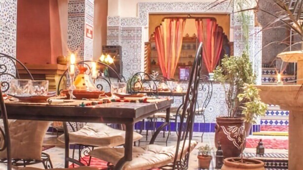 A dining area with a table set for two, surrounded by chairs, and a decorative fountain in the background. - Riad Mahjouba Marrakech & SPA (Marrakech)