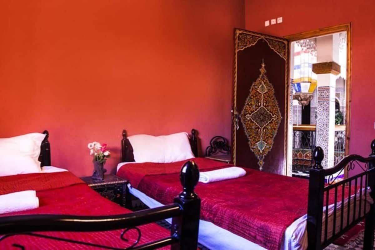 A room with two beds, each with a red bedspread and white pillows. There is a decorative door with intricate patterns and a small table with a vase of flowers.
