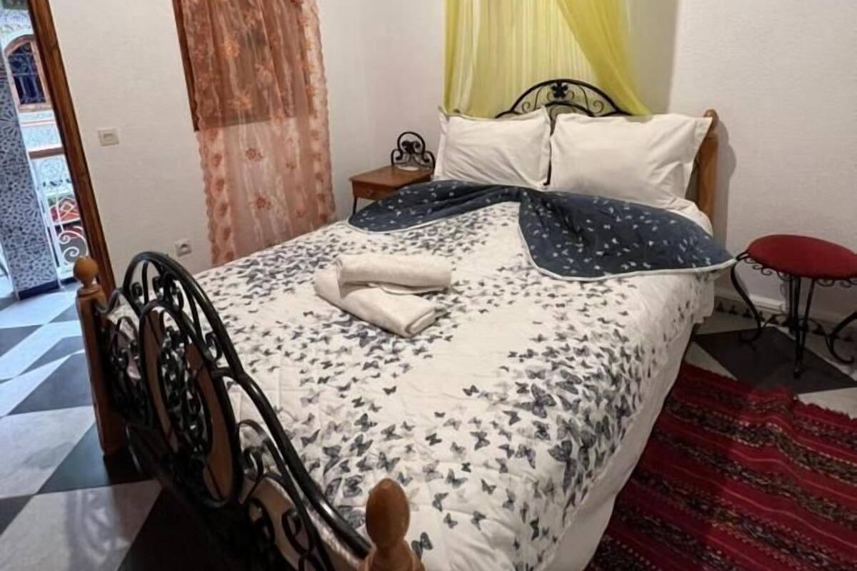 A bed with a canopy, a bedside table, a red stool, and a patterned rug.