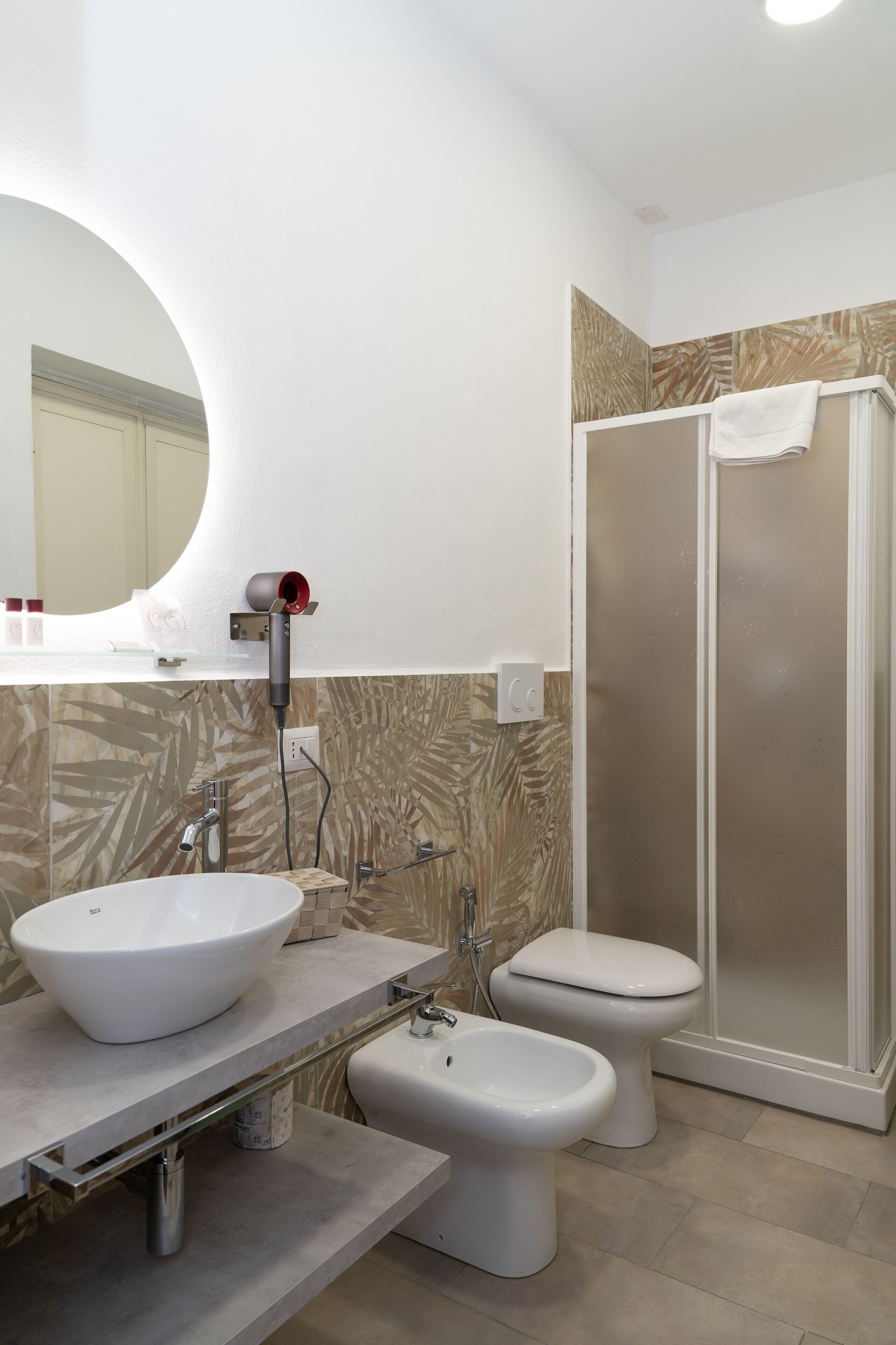 A modern bathroom with a large oval mirror, a white sink, a bidet, a toilet, and a walk-in shower.