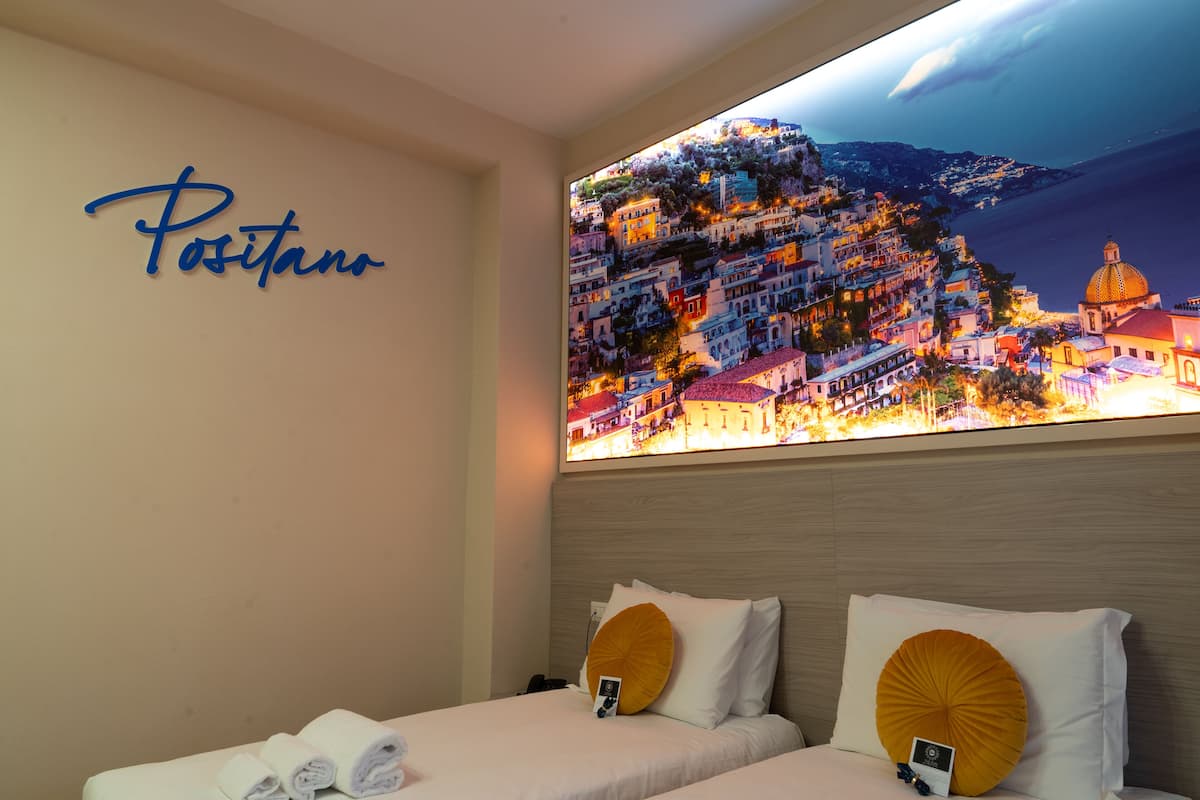 A hotel room with a bed, a wall-mounted artwork of a coastal town, and a sign reading "Positano".