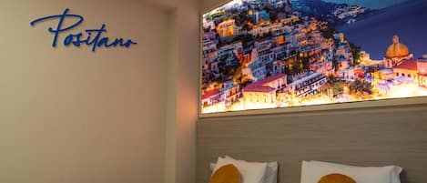 A hotel room with a bed, a wall-mounted artwork of a coastal town, and a sign reading "Positano".