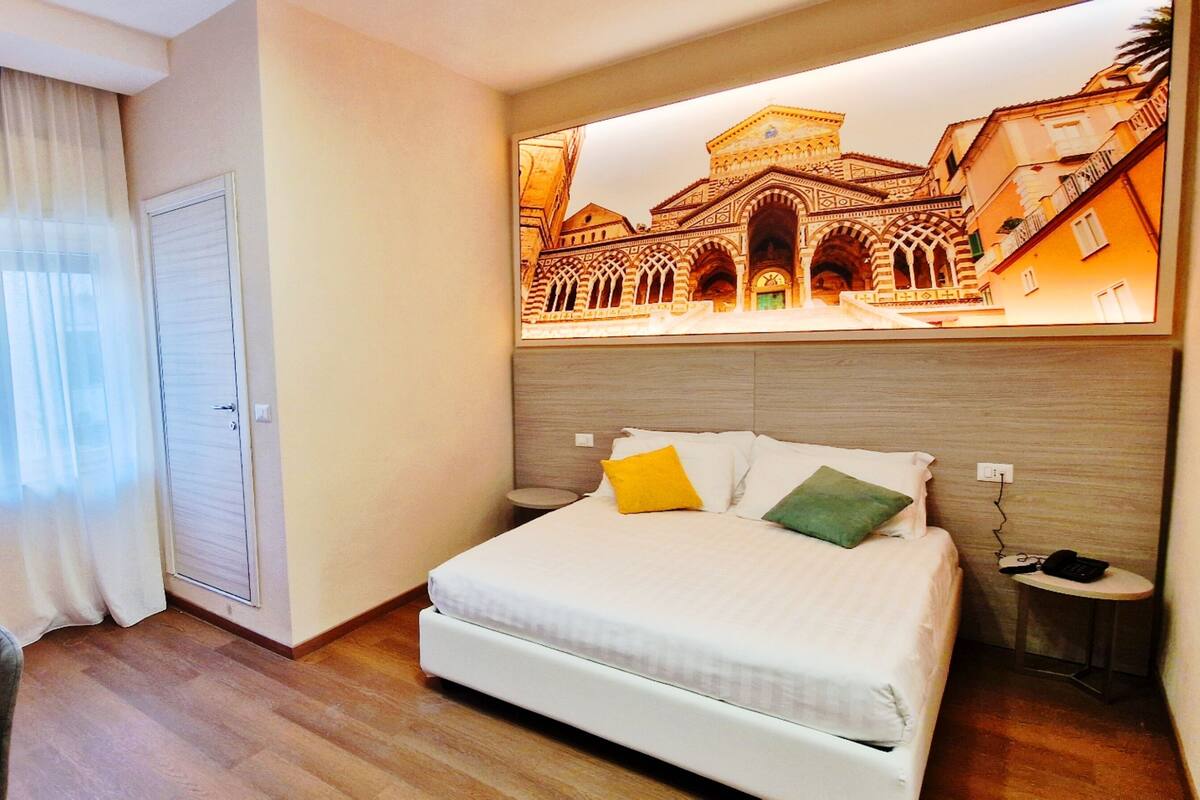 A hotel room with a bed, a desk, a chair, a window with curtains, and a wall-mounted artwork of a building.