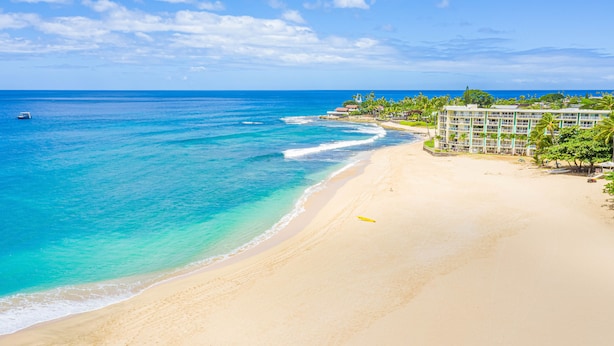 Una playa con aguas turquesas cristalinas, una orilla arenosa y un edificio de resort con varios pisos y balcones con vista al océano.