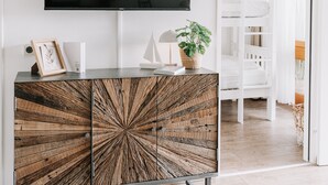 Un salon moderne avec un grand téléviseur à écran plat fixé à un mur blanc, un meuble en bois au design rayonnant, une petite plante en pot et, au fond, un lit superposé blanc.
