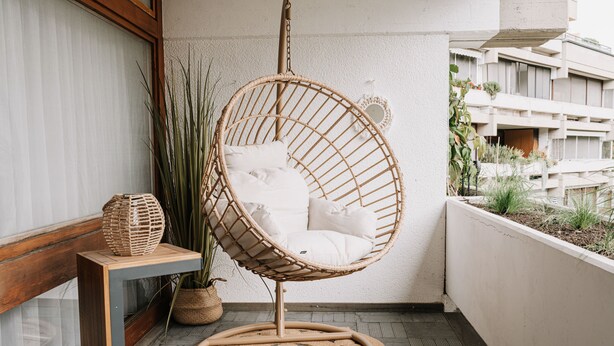 Un fauteuil suspendu en osier, doté de coussins blancs, se trouve sur un balcon dont le sol est en bois. On y aperçoit également une petite table sur laquelle repose un panier tressé, ainsi qu’une plante en pot.