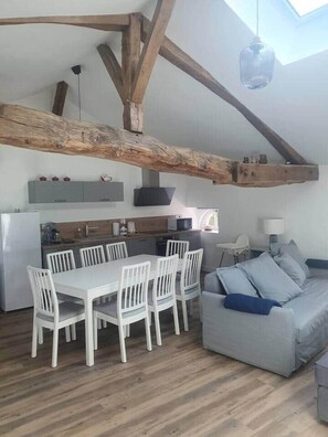 Une cuisine moderne avec des poutres en bois, un coin repas avec des chaises blanches et un canapé gris.