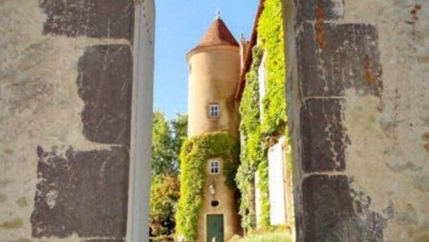 An arched stone doorway leading to a tower with a red roof, surrounded by greenery.
