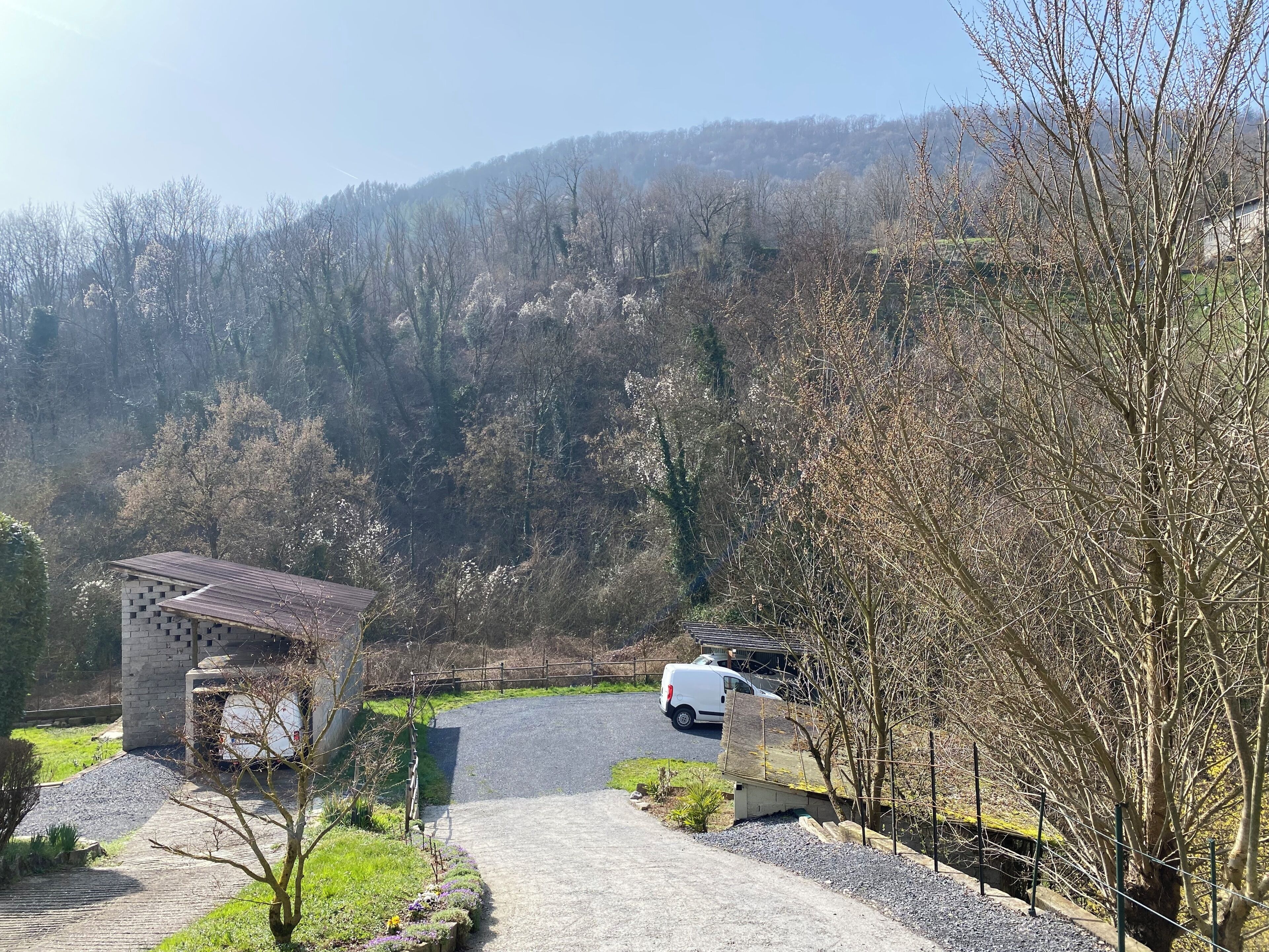 A paved driveway leading to a building with a brick facade, surrounded by trees and a hilly landscape.