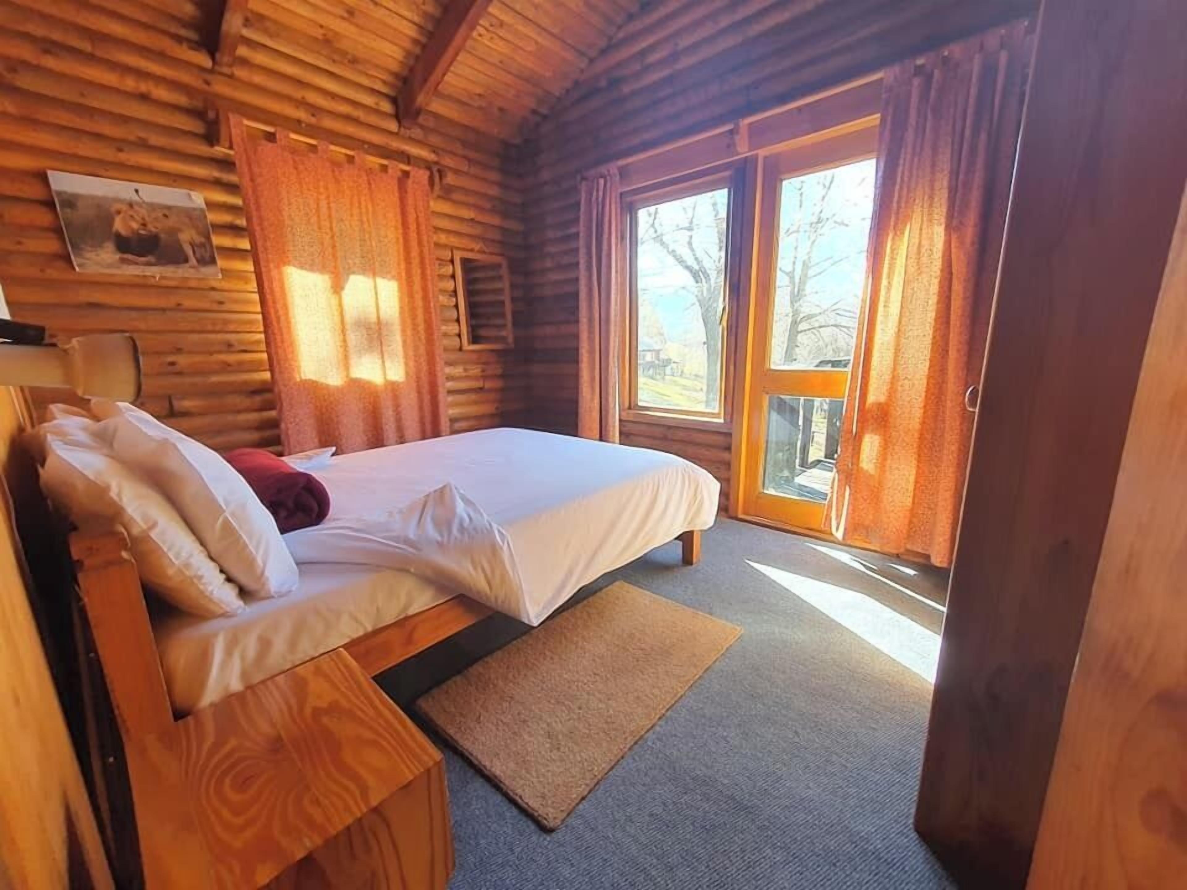 A wooden cabin bedroom with a bed, pillows, a wooden nightstand, a window with curtains, and a door leading outside.