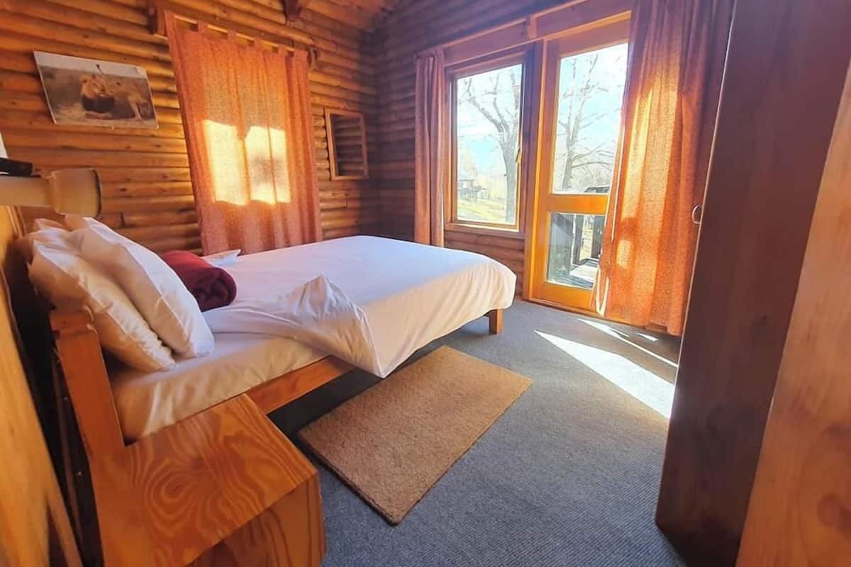 Un dormitorio de cabaña de madera con una cama, almohadas, una mesita de noche de madera, una ventana con cortinas y una puerta que da al exterior.