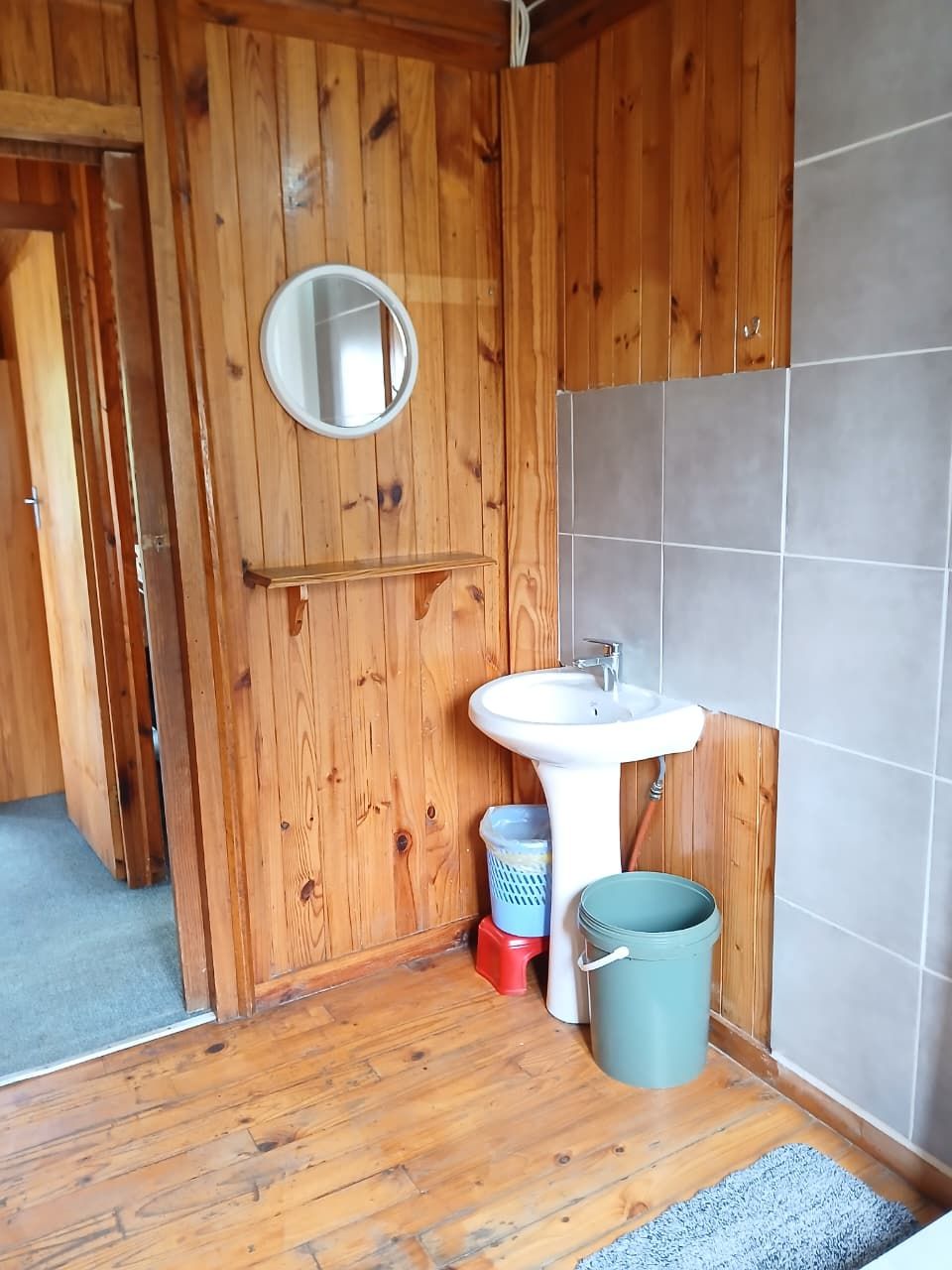 A wooden-paneled bathroom with a white sink, a round mirror, and a grey bucket.