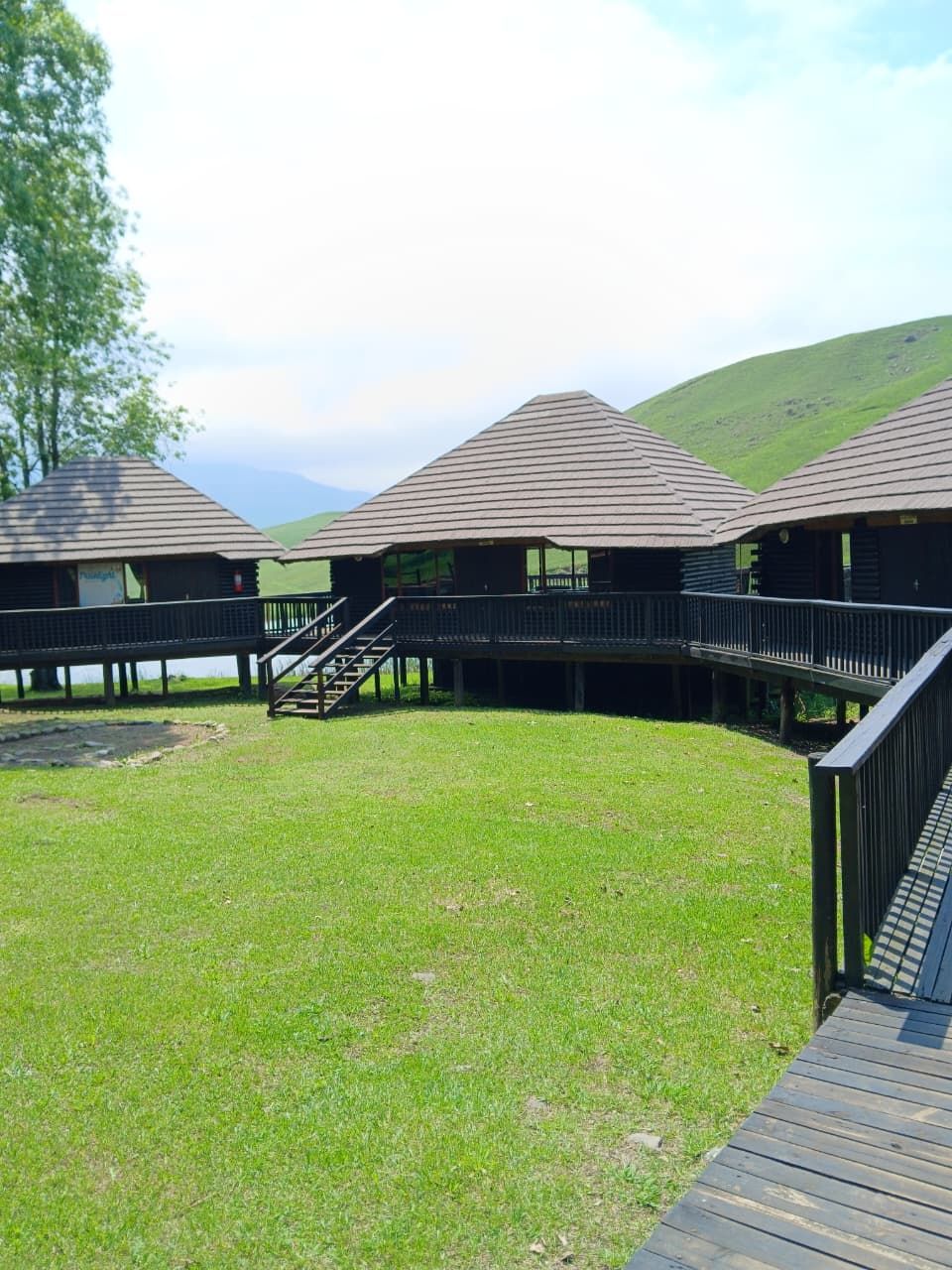 Várias cabanas de madeira com varandas, unidas por um deck de madeira, com colinas e céu claro ao fundo.