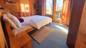 Ein Schlafzimmer in einer Holzhütte mit einem Bett, Kissen, einem Nachttisch aus Holz, einem Fenster mit Vorhängen und einer Tür, die nach draußen führt.
