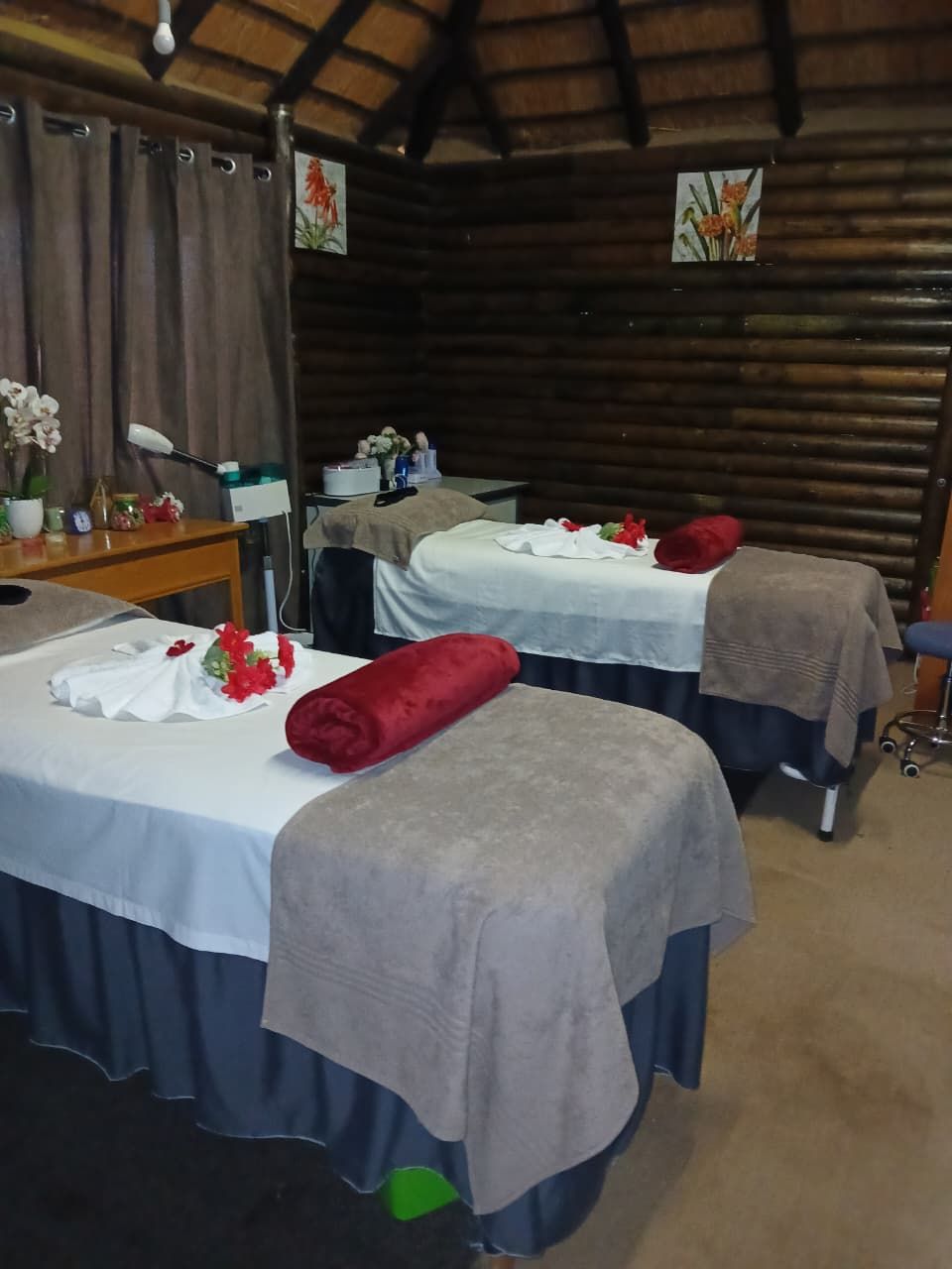Habitación con dos camas, cada una con sábana blanca y manta marrón, y una mesita con lámpara y flores.